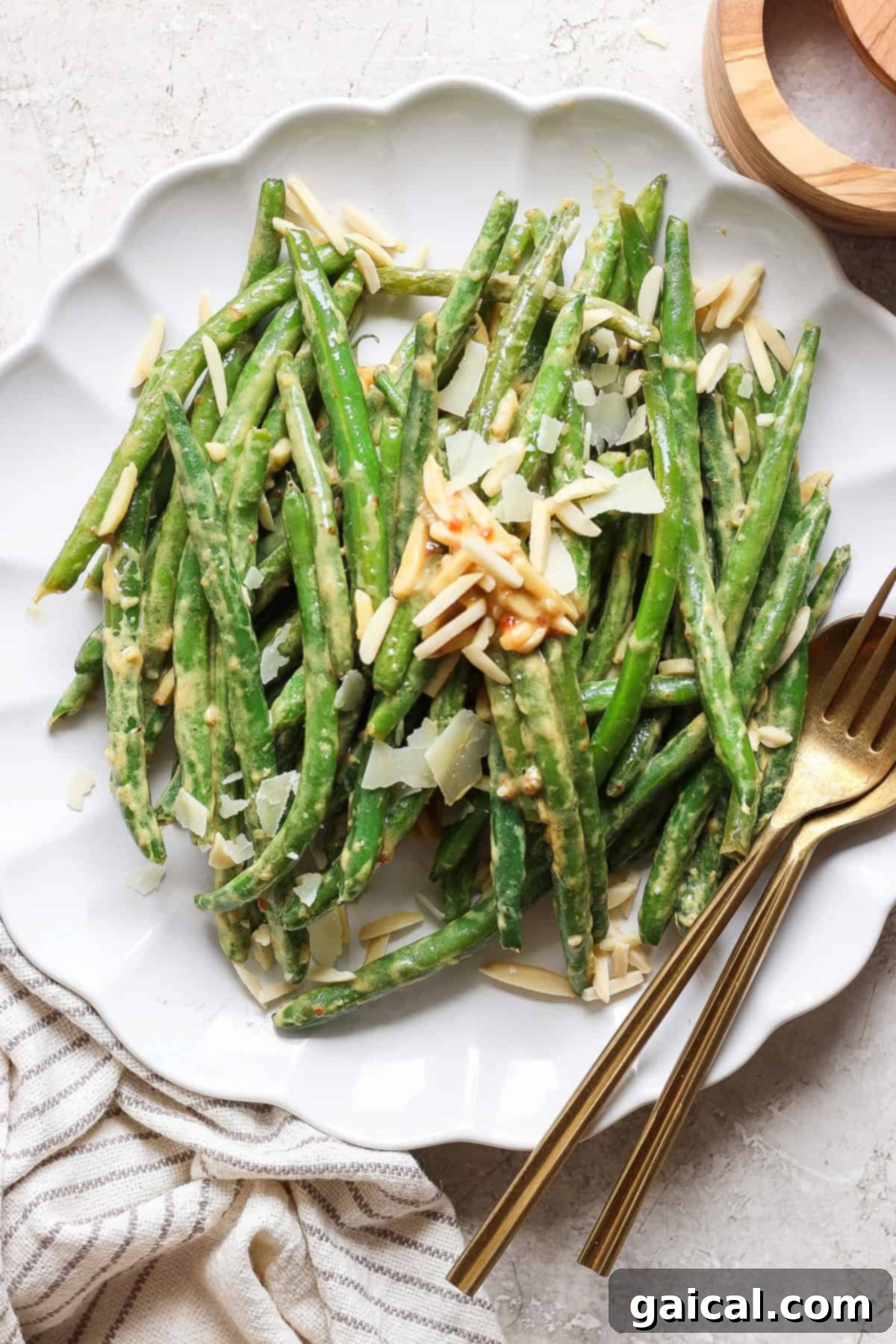 A beautifully plated serving of vibrant green beans, generously topped with golden slivered almonds and delicate shavings of Parmesan cheese. The dish rests on a pristine white tablecloth, accompanied by elegant gold cutlery, inviting a delightful culinary experience.