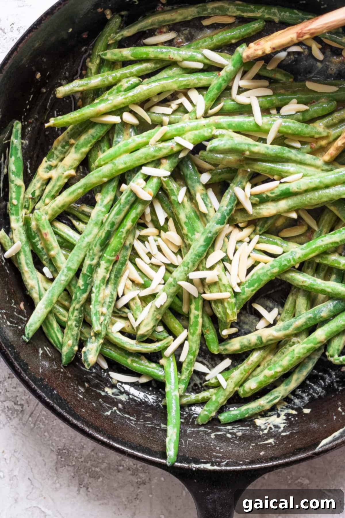 A close-up shot of a black cast iron skillet filled with perfectly cooked green beans, glistening with a creamy sauce and generously garnished with slivered almonds. A wooden spoon rests gently on the rim, ready for serving.
