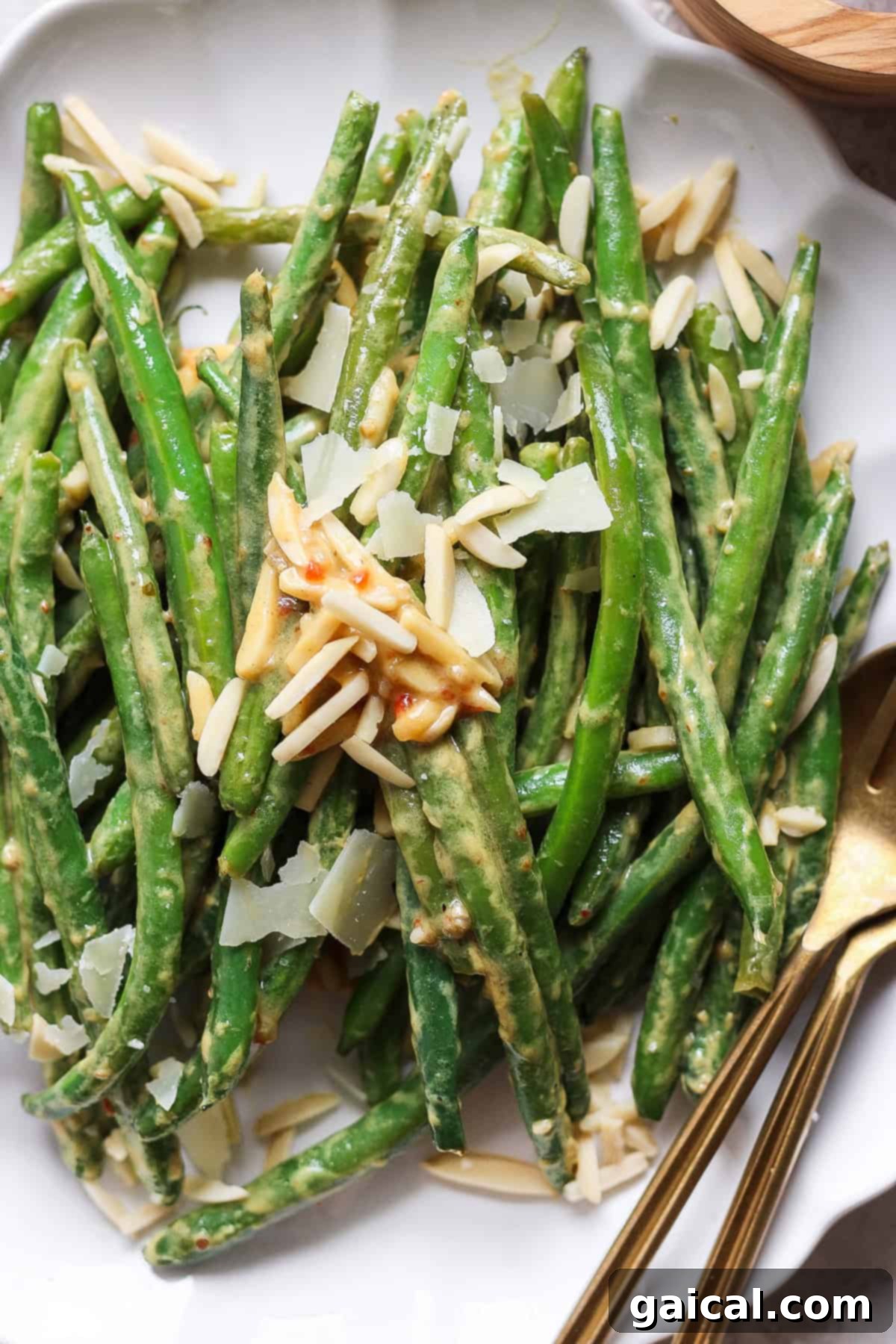 A delightful plate of vibrant green beans elegantly topped with fluffy shaved Parmesan cheese and crunchy almond slivers, presented invitingly with serving utensils placed alongside the dish, ready to be enjoyed.