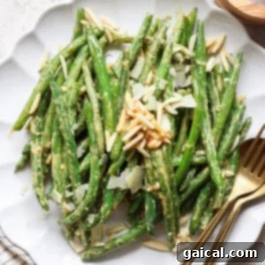 A plate of perfectly cooked green beans, elegantly topped with fluffy shaved Parmesan cheese and crunchy almond slivers, presented invitingly with serving utensils.