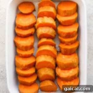 A white baking dish filled with sliced sweet potatoes arranged in three rows on a light surface.