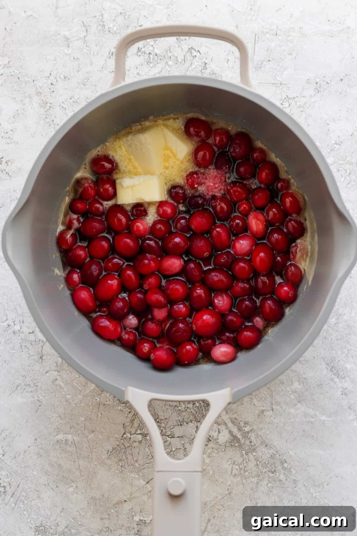 Golden Harvest Maple Cranberry Sweet Potatoes 5 A gray pot contains raw cranberries, a block of butter, and sugar on a light textured surface.
