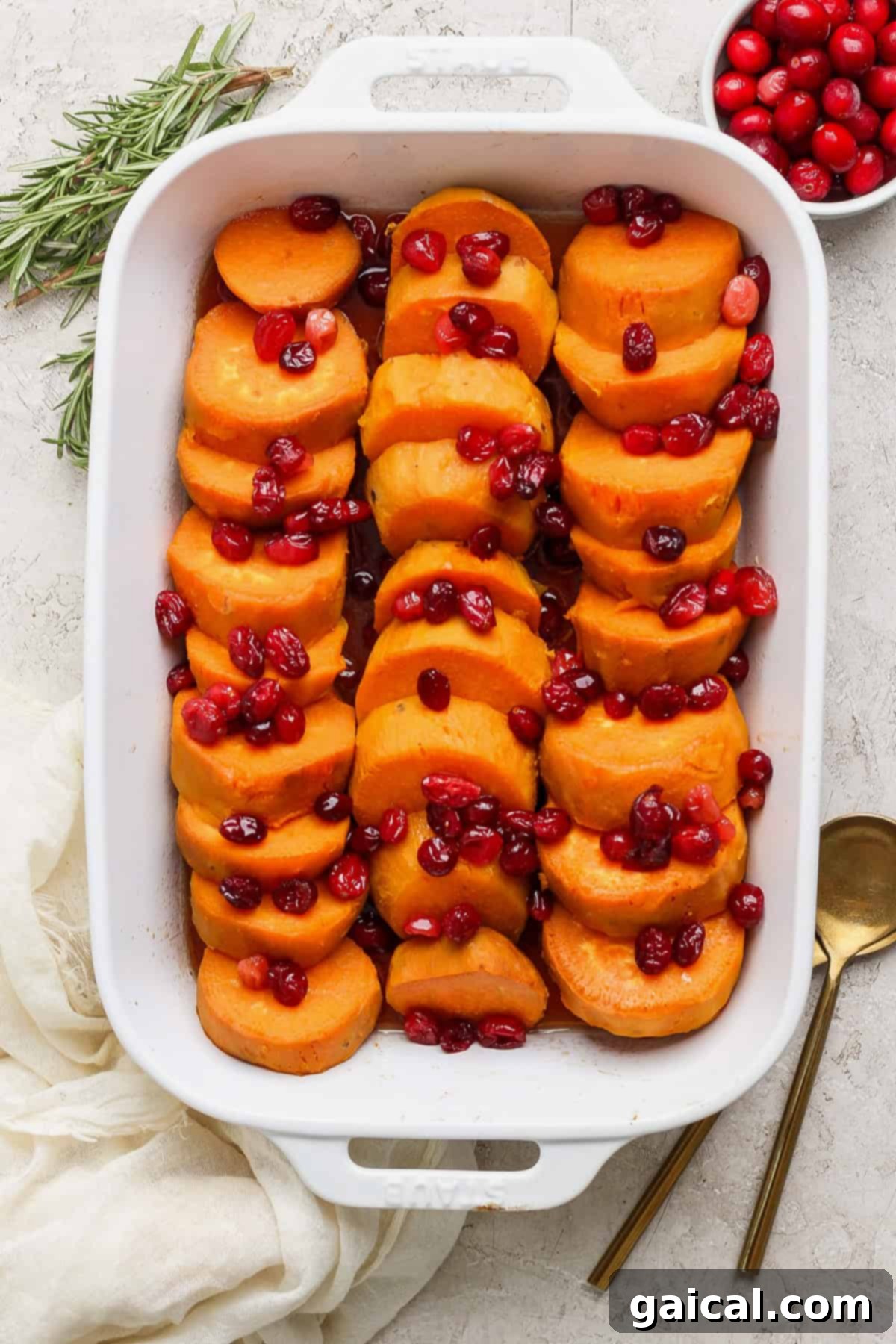 Golden Harvest Maple Cranberry Sweet Potatoes 7 A white baking dish filled with sliced sweet potatoes topped with cranberries. A sprig of rosemary, a bowl of cranberries, and two gold utensils are placed beside the dish.