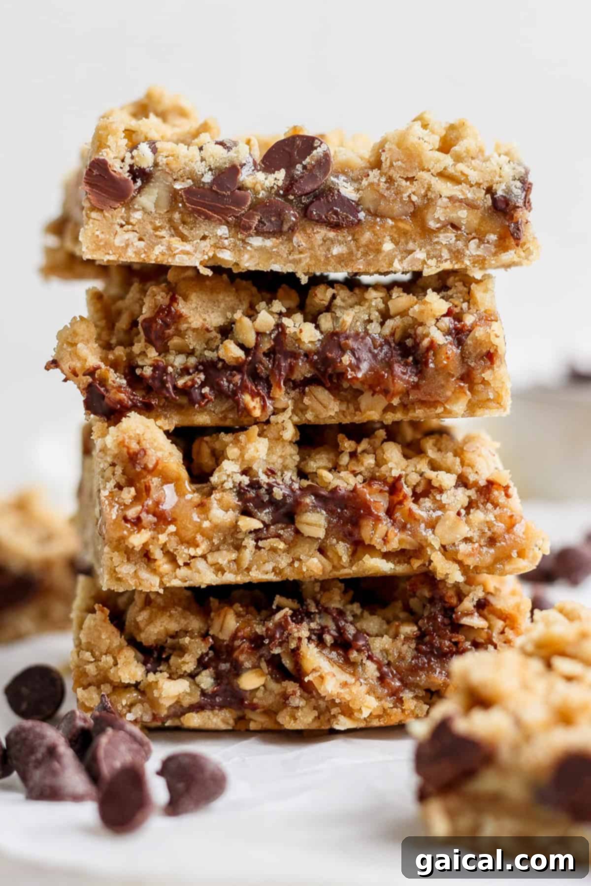 Nana Mabels Carmelita Bars 2 A beautifully stacked set of four homemade Carmelita bars, featuring visible layers of oats, chocolate, and caramel, presented on a pristine white surface with scattered chocolate chips for garnish.