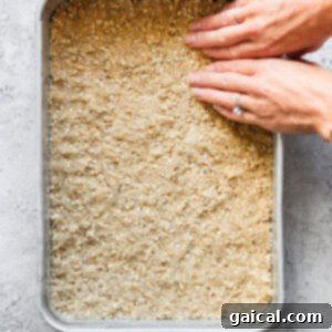 A pair of hands gently but firmly pressing down a golden-brown, crumbly mixture, visibly rich with oats, into an even and compact layer within a square baking pan. The mixture is spread uniformly across the pan's base.