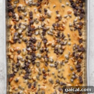 A metal baking tray generously filled with a layered dessert, beautifully topped with a shimmering layer of melted caramel, an abundance of sweet chocolate chips, and crunchy chopped nuts, prepared for baking.