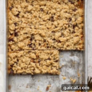 A metal baking pan filled with golden-brown, crumbly dessert bars. Two bars have been neatly cut out and removed, revealing a delicious layered filling, while the rest of the dessert remains enticingly uncut. A small fork and a white napkin are placed invitingly nearby.