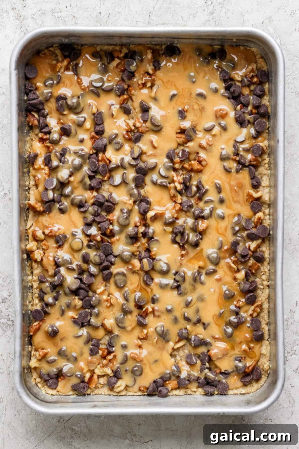 Nana Mabels Carmelita Bars 4 A gleaming metal baking tray showcasing an unbaked dessert, meticulously topped with a generous layer of melted caramel, an abundance of rich chocolate chips, and finely chopped nuts, ready for its final bake.