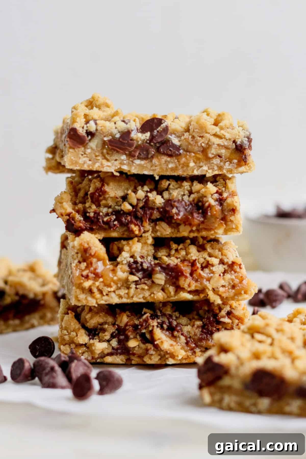 Nana Mabels Carmelita Bars 6 A perfectly arranged stack of four rich chocolate chip oatmeal bars, featuring a tempting crumbly topping, displayed elegantly on a clean white surface. A scattering of additional chocolate chips surrounds the base of the stack, enhancing its visual appeal.