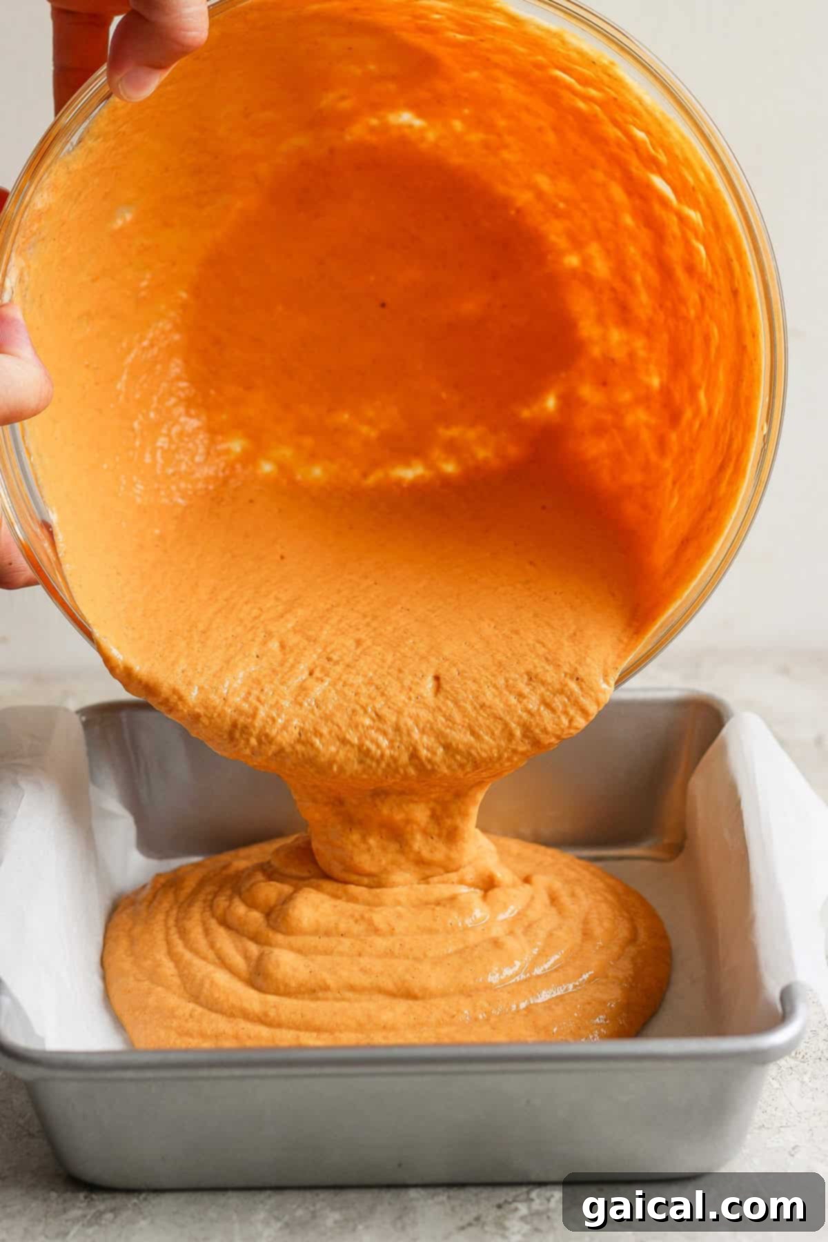 Hands pouring orange batter from a bowl into a parchment-lined baking tin.