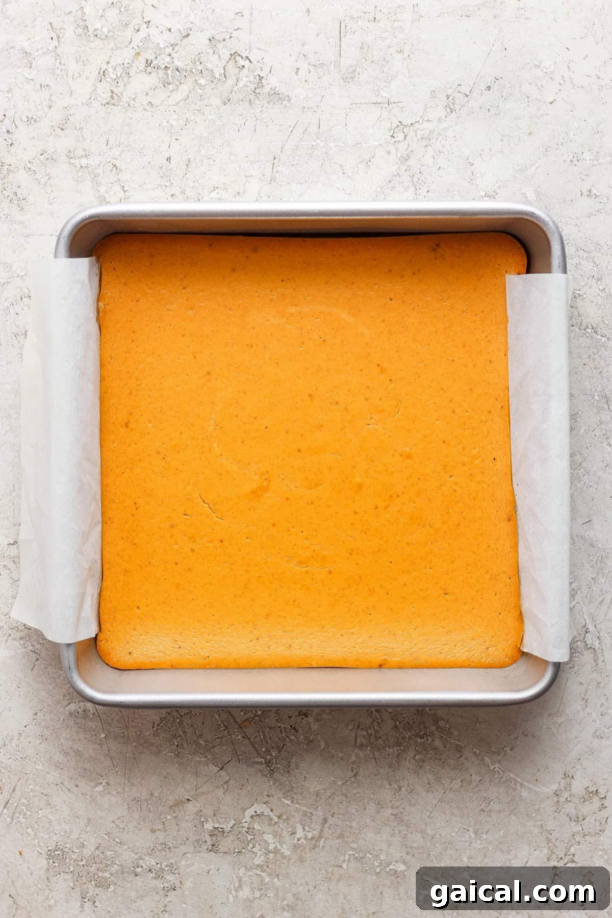 A square pan with a light golden-brown baked cake lined with parchment paper on a textured, light-colored surface.