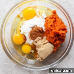 A glass mixing bowl containing raw eggs, sour cream, pumpkin puree, brown sugar, and spices, ready to be mixed.