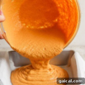 Hands pouring orange batter from a bowl into a parchment-lined baking tin.