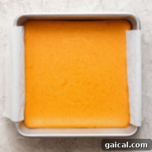 A square pan with a light golden-brown baked cake lined with parchment paper on a textured, light-colored surface.