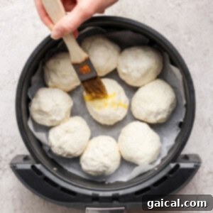 A hand brushes egg wash on dough balls arranged in an air fryer basket lined with parchment paper.