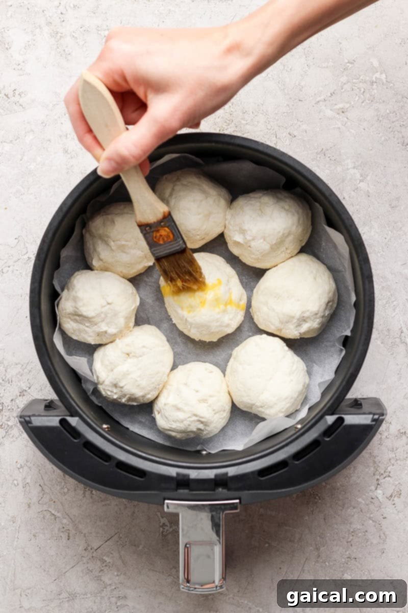 A hand brushes egg wash on dough balls arranged in an air fryer basket lined with parchment paper.