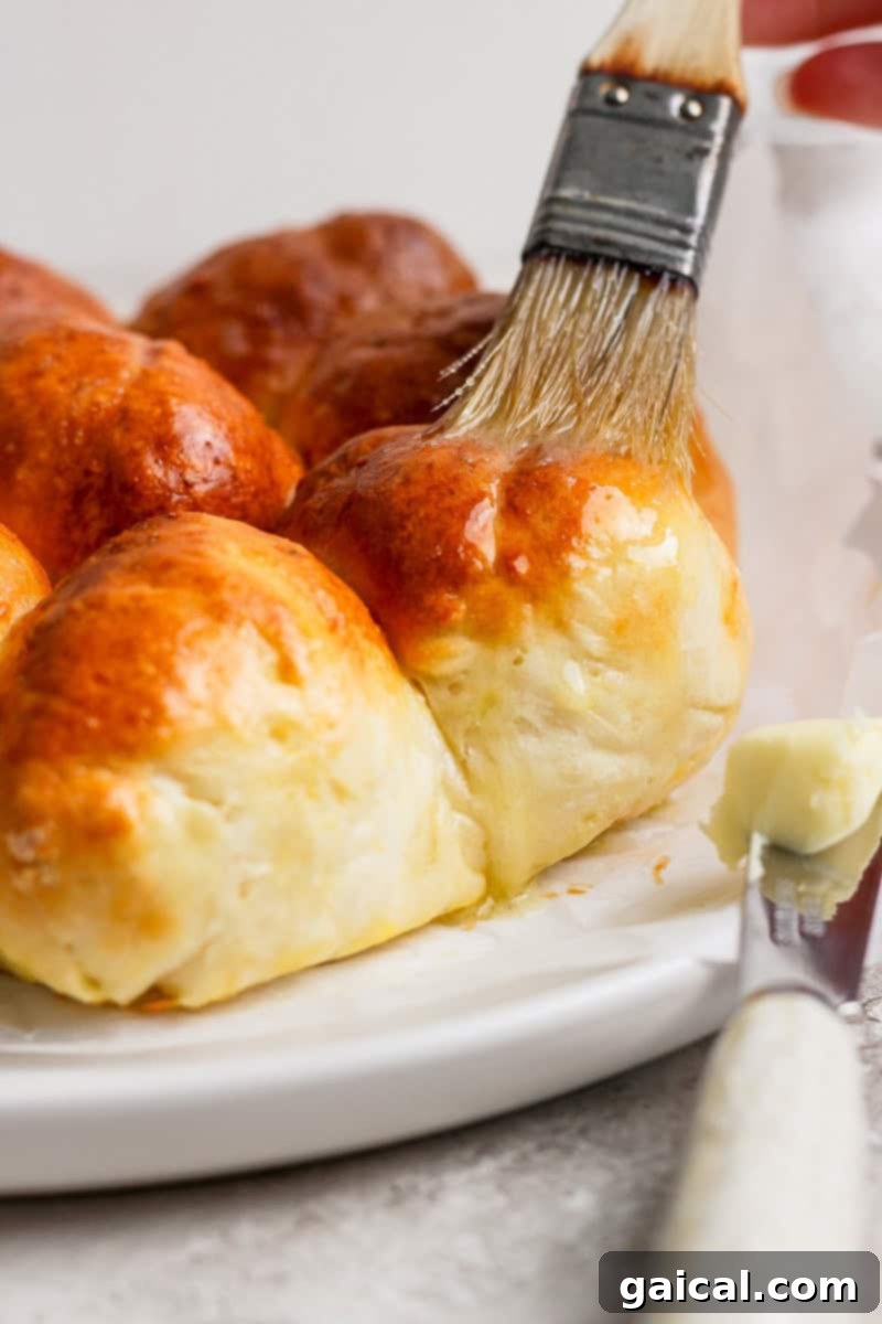 A pastry brush applies butter to freshly baked golden-brown bread rolls on a white plate.