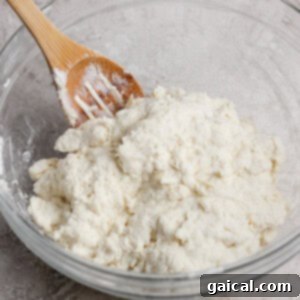 A wooden spoon rests in a glass bowl filled with a mixture of flour and a white ingredient, likely dough, on a textured countertop.