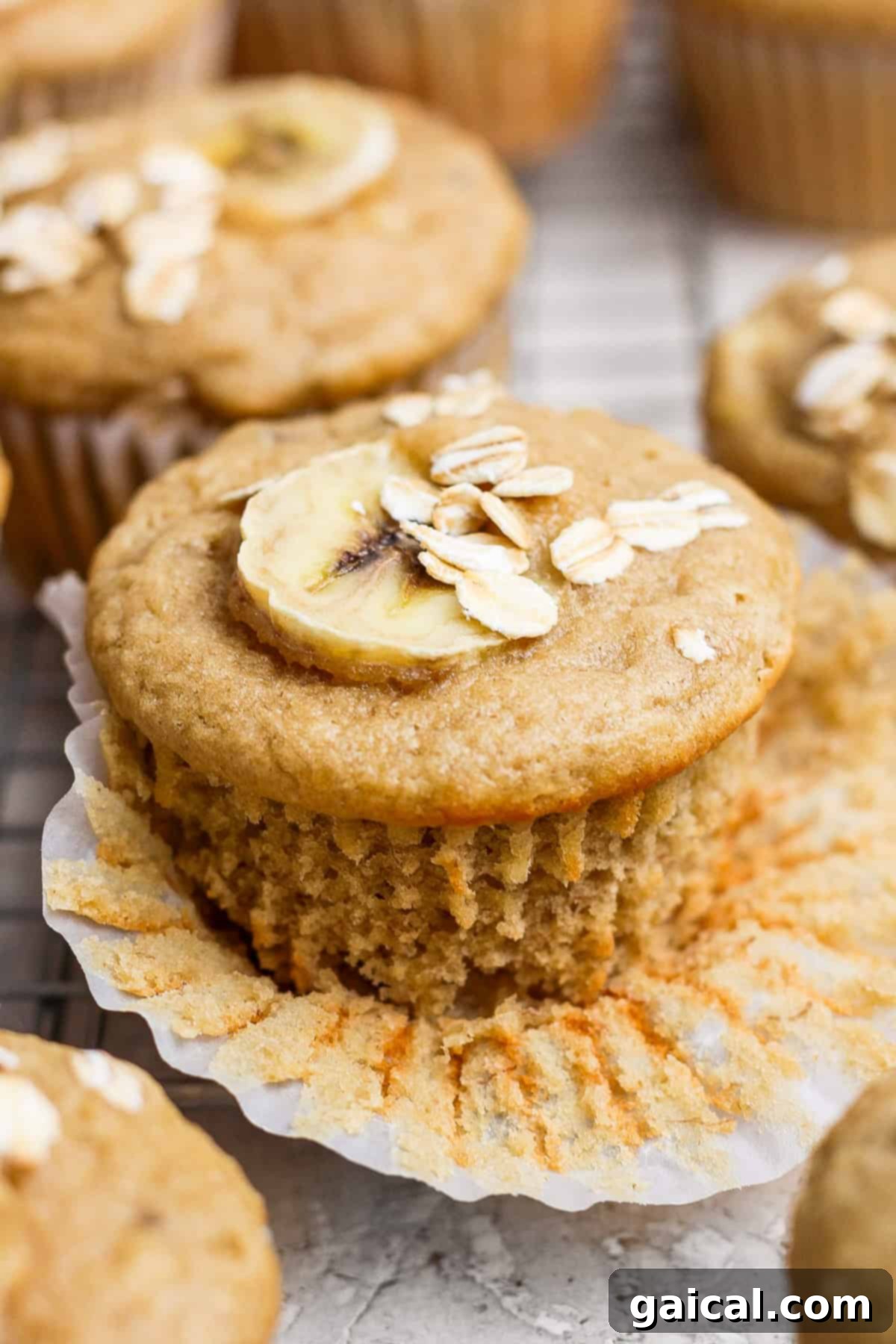 Wholesome Banana Oat Muffins 2 A beautifully baked banana muffin, topped with a delicate banana slice and a sprinkle of oats, gently unwrapped on a cooling wire rack.