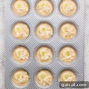 A muffin tray with twelve muffin batter portions, each topped with banana slices and oats, perfectly portioned and ready for baking.