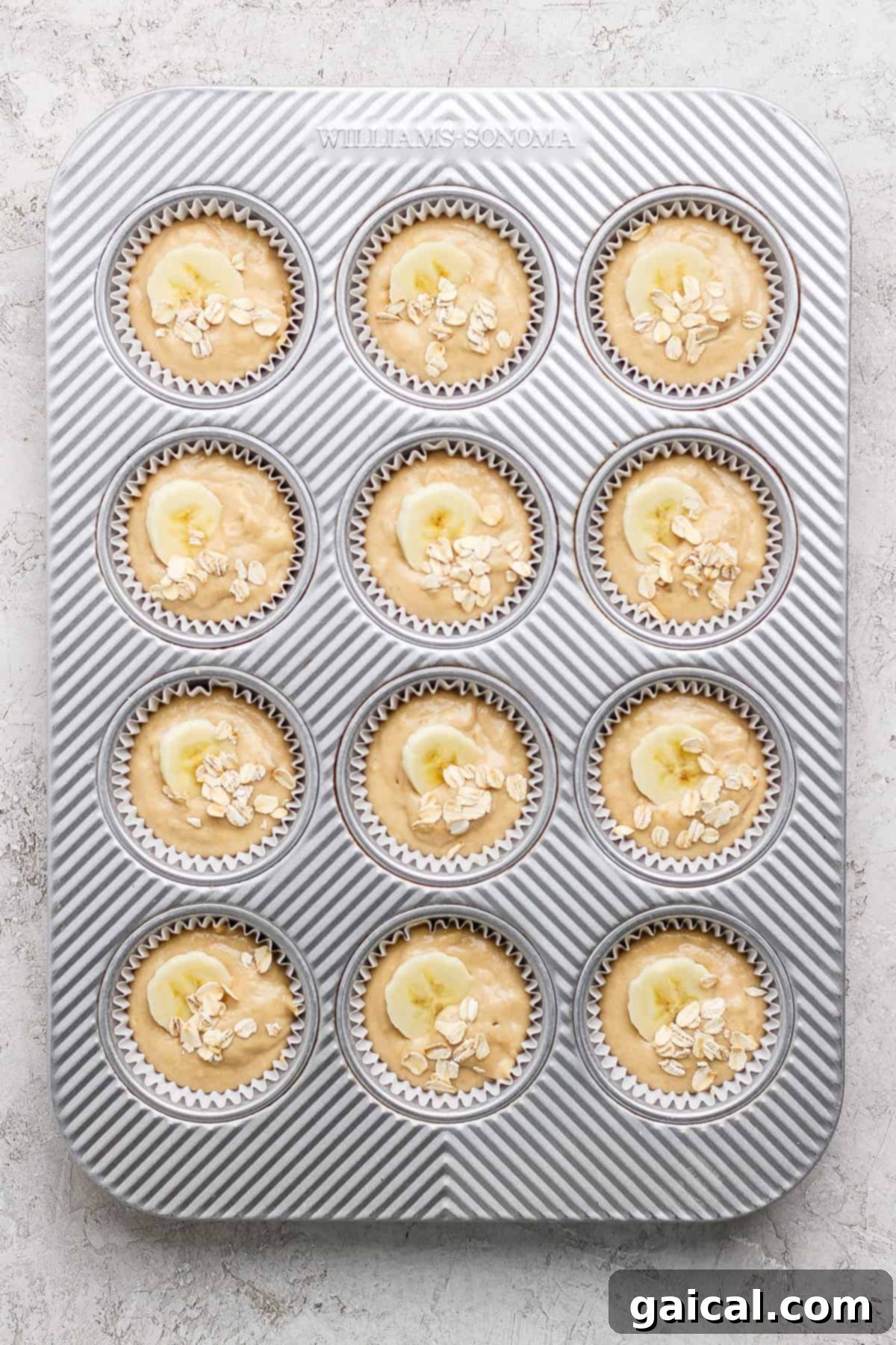 Wholesome Banana Oat Muffins 4 A muffin tray filled with twelve portions of muffin batter, each beautifully topped with thin banana slices and a sprinkle of oats, ready for baking.