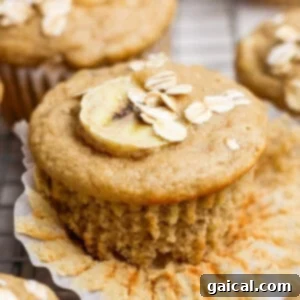 A banana muffin topped with a banana slice and oats, with a paper liner partially peeled back, ready to eat.