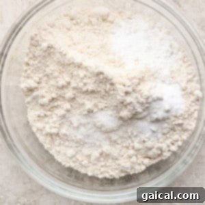 A clear glass bowl containing a mixture of flour and baking powder on a textured beige surface, ready for whisking.