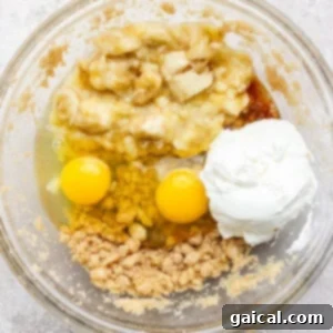 A glass bowl containing mashed bananas, two eggs, yogurt, brown sugar, and liquid coconut oil, ready to be mixed for muffin batter.