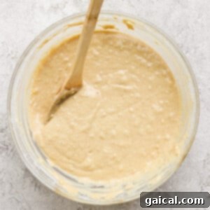 A glass bowl filled with creamy muffin batter and a wooden spoon resting in it on a textured white surface, showing the perfect consistency.