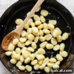 A cast iron skillet filled with cooked gnocchi, stirred with a wooden spoon, on a light gray textured surface.