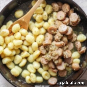 A skillet containing cooked gnocchi and sliced sausages mixed with a sauce, with a wooden spoon resting inside.