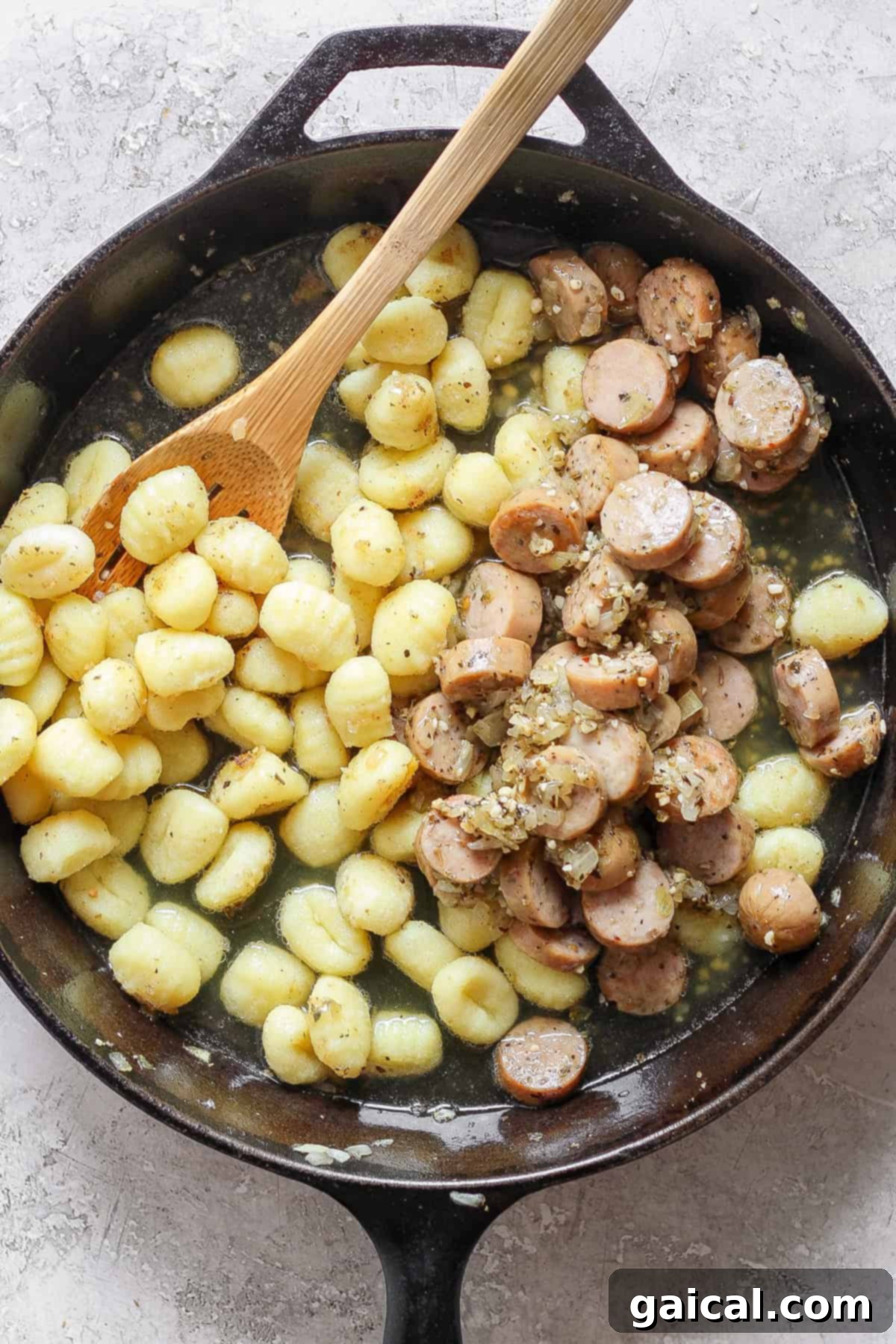 A skillet containing cooked gnocchi and sliced sausages mixed with a sauce, with a wooden spoon resting inside.