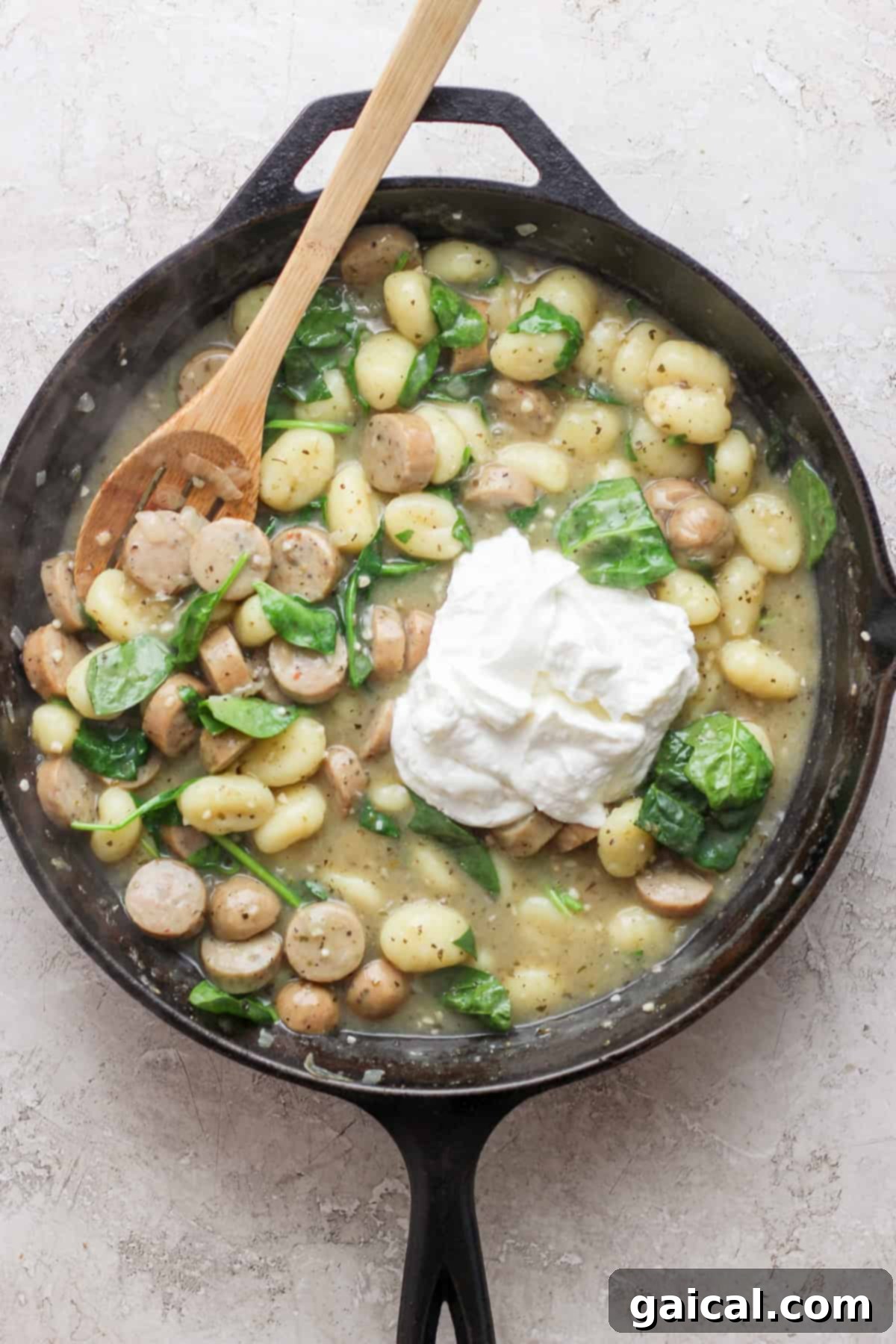 A skillet filled with gnocchi, spinach, sliced sausage, and a dollop of ricotta cheese, with a wooden spoon rested on the edge.