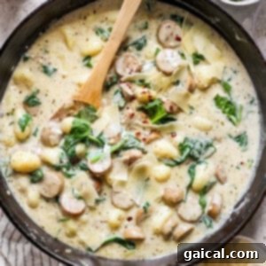 A skillet filled with creamy gnocchi, spinach, and sausage topped with shredded cheese and red pepper flakes. A wooden spoon rests inside the skillet.