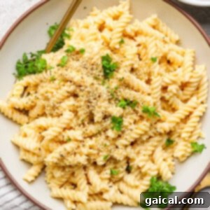 A bowl of rotini pasta with creamy sauce, topped with chopped herbs and breadcrumbs, on a round plate with a fork.