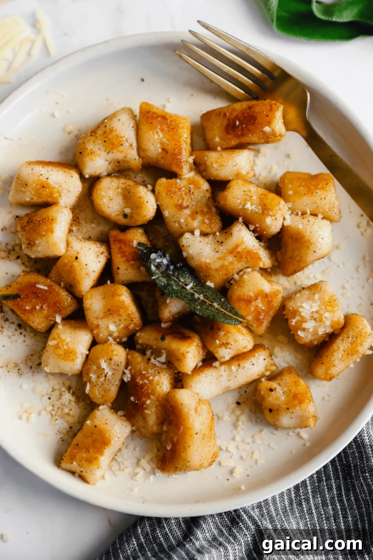 Pillowy Cottage Cheese Gnocchi 2 Plate of golden brown cottage cheese gnocchi with grated cheese and a sage leaf, accompanied by a fork, on a white background.