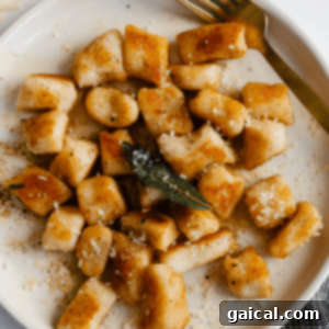 A plate of golden-brown cottage cheese gnocchi topped with fried sage and grated cheese, accompanied by a gold fork.
