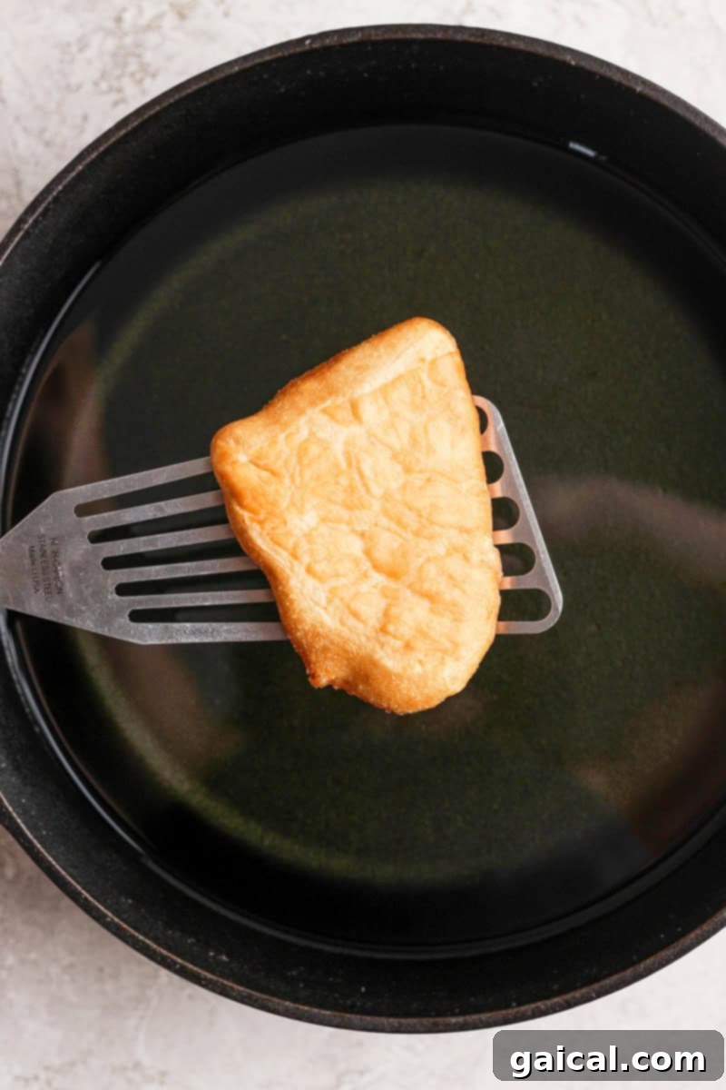 Nonna Marie's Crispy Fried Pizza 5 A piece of golden-brown fried bread being carefully lifted from a pan of shimmering hot oil with a metal spatula, steam gently rising.