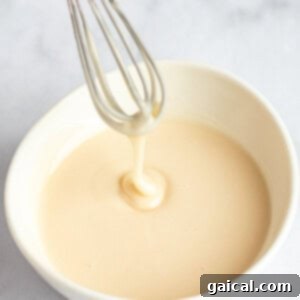 Fueling Frosted Pastries 14 A whisk with creamy batter drips into a white bowl on a light surface.