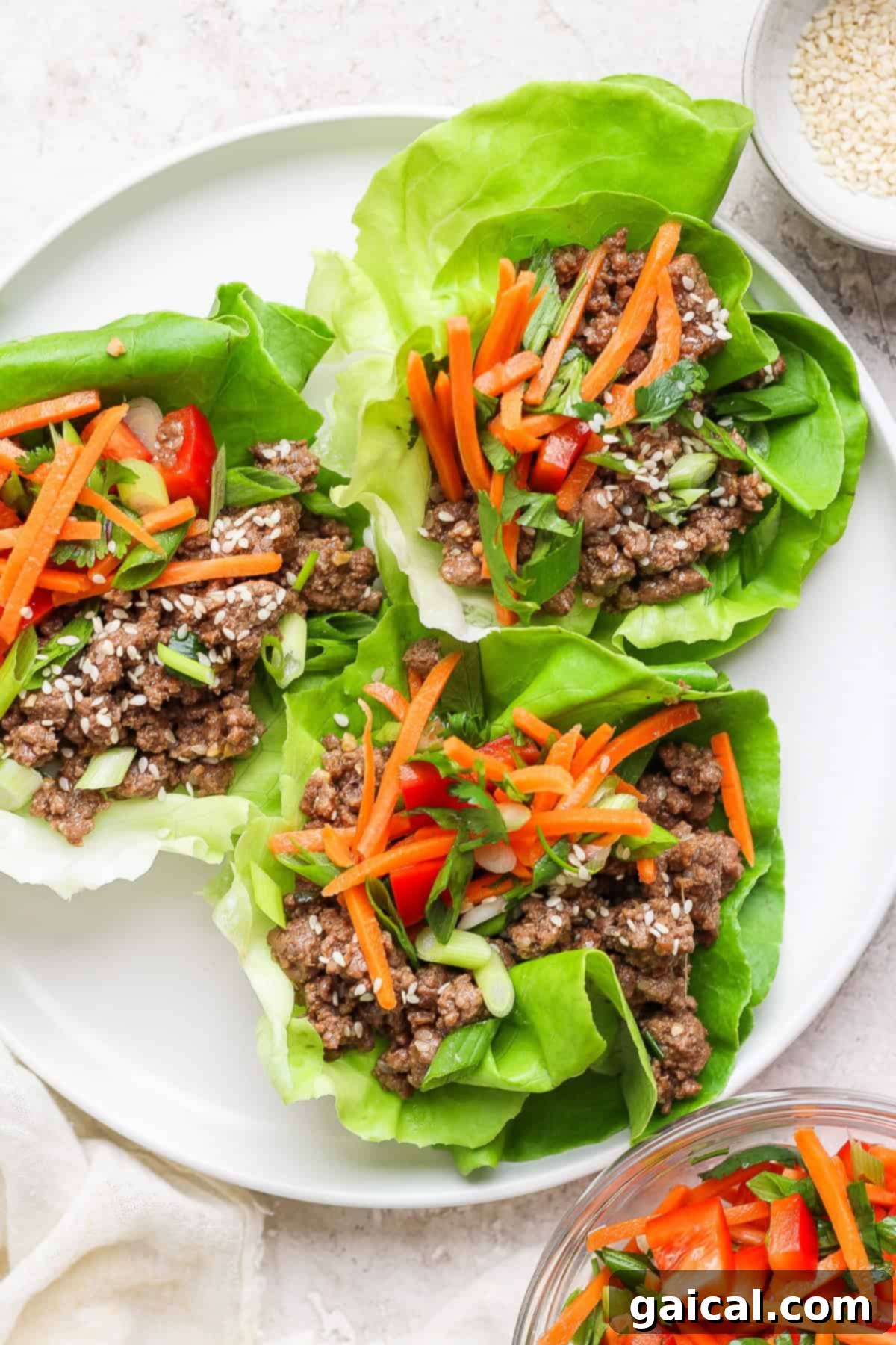 Four crisp lettuce wraps filled with succulent Korean ground beef, vibrant shredded carrots, finely diced red bell peppers, and fresh green onions, elegantly arranged on a pristine white plate.