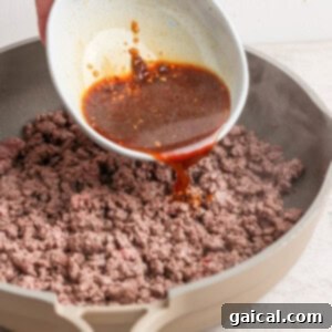 A hand pours a brown sauce from a white bowl onto cooked ground beef in a beige skillet.