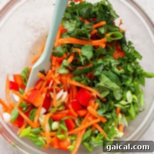 A glass bowl filled with chopped vegetables, including red bell peppers, carrots, green onions, cilantro, and green bell peppers, with a light blue spatula.