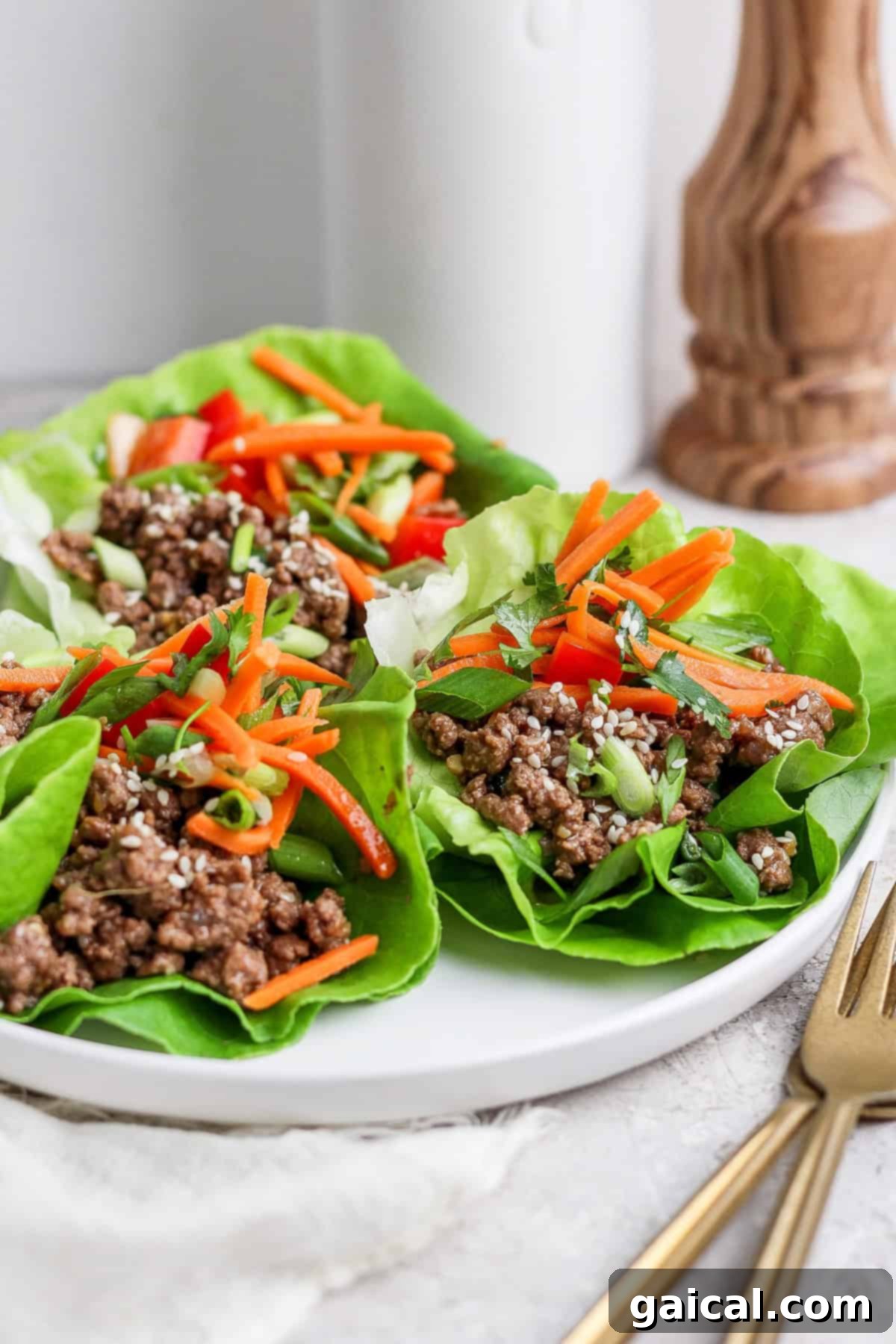 Exquisitely presented lettuce wraps, brimming with seasoned minced meat, thinly sliced carrots, a sprinkle of sesame seeds, and vibrant green onions, served on a white plate alongside elegant gold cutlery.