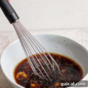 A whisk is stirring a dark-colored sauce in a light gray bowl on a textured surface.
