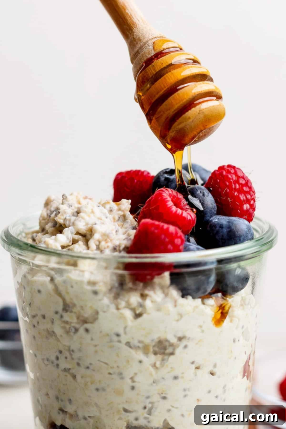 A glass jar of cheesecake-inspired overnight oats, generously topped with vibrant blueberries and raspberries, and finished with a delicate drizzle of honey from a wooden dipper. Photographed on a rustic wooden surface.