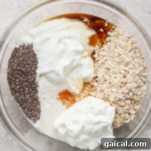 A clear bowl containing cheesecake overnight oats, yogurt, chia seeds, honey, and milk rests on a textured surface.