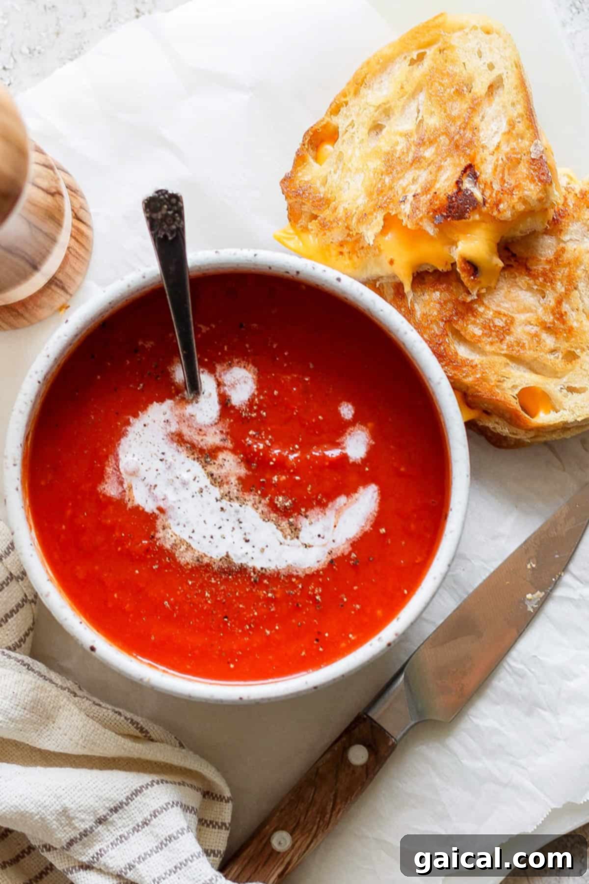 A bowl of tomato soup swirled with cream and pepper sits beside a grilled cheese sandwich, accompanied by a knife and a striped napkin.