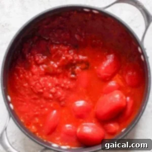 A black pot filled with whole and crushed tomatoes in tomato sauce, reminiscent of a comforting bowl of tomato soup, sits on a light gray surface. Perfect for pairing with a crispy grilled cheese sandwich.