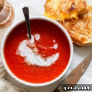 A bowl of tomato soup swirled with cream and pepper sits beside a grilled cheese sandwich, accompanied by a knife and a striped napkin.