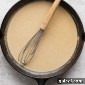 A cast iron skillet filled with a light brown, creamy sauce, with a whisk resting inside, signaling the sauce is almost ready.